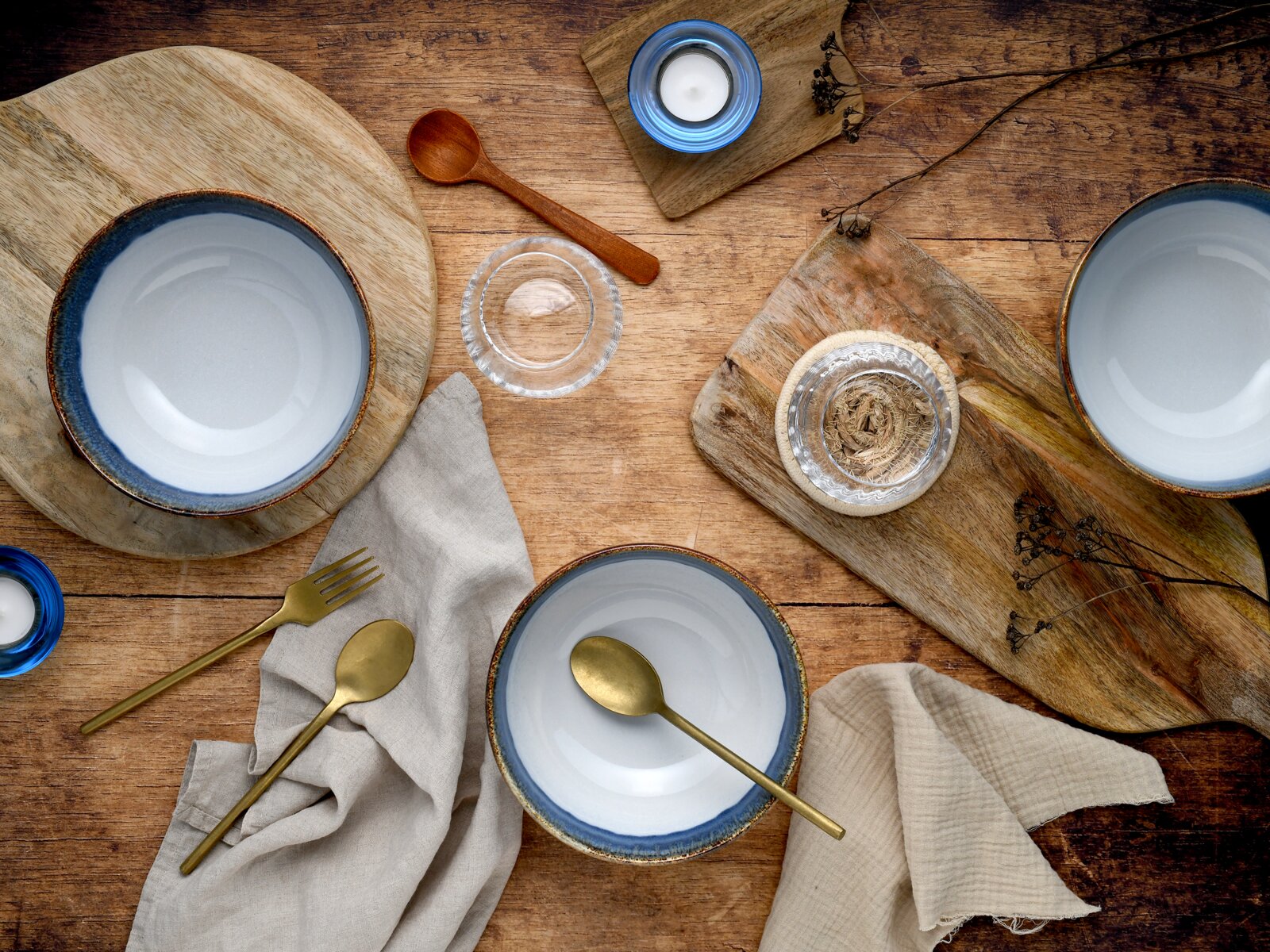Rustikales Tisch-Arrangement aus Keramikschalen mit blauem Rand, goldfarbenem Besteck, Holzbrettchen und Kerzen auf Holztisch, aufgenommen aus der Vogelperspektive (Draufsicht).