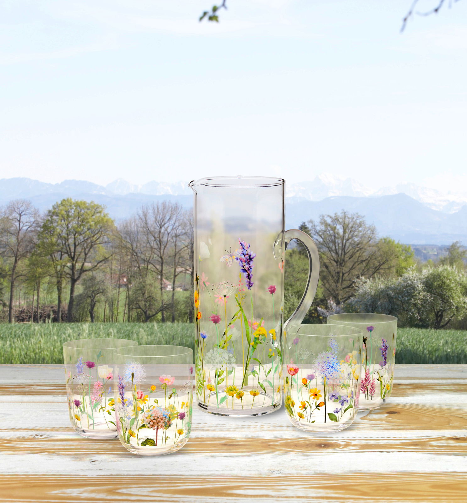 Gläserset mit Blumenmuster, bestehend aus einem Krug und vier Gläsern, auf einem Holztisch mit Blick auf eine Landschaft mit Bäumen und Bergen im Hintergrund.