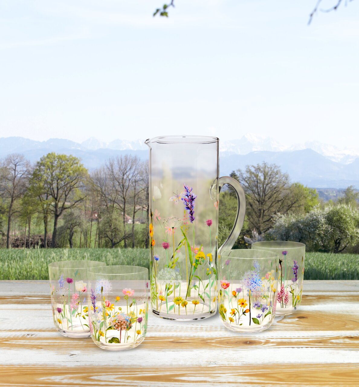 Gläserset mit Blumenmuster, bestehend aus einem Krug und vier Gläsern, auf einem Holztisch mit Blick auf eine Landschaft mit Bäumen und Bergen im Hintergrund.