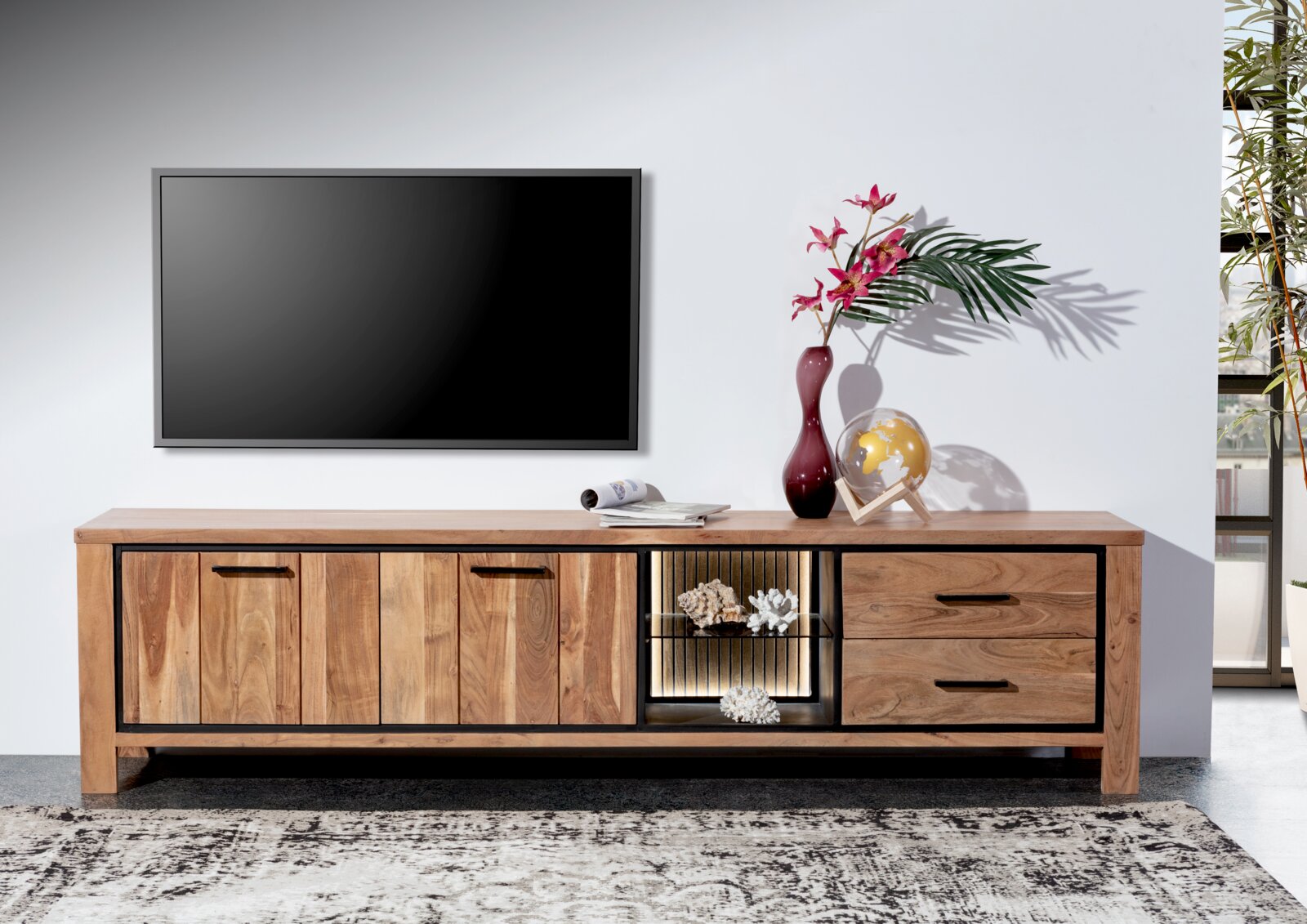 Frontale Ansicht eines Lowboards aus Holz mit zwei Türen, zwei Schubladen und einem offenen Fach mit Beleuchtung, dekoriert mit einer Vase, einem Globus und Zeitschriften.