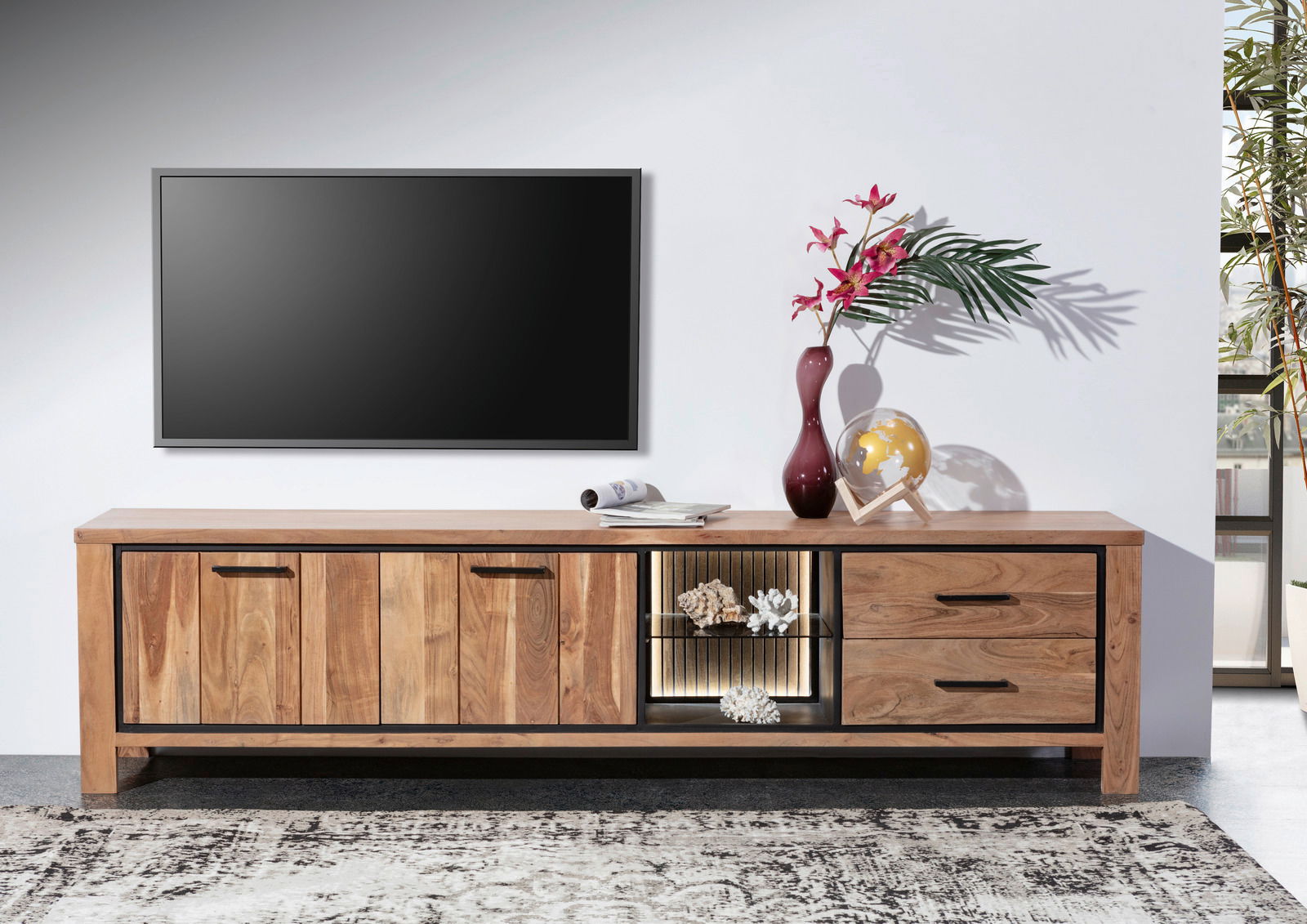 Frontale Ansicht eines Lowboards aus Holz mit zwei Türen, zwei Schubladen und einem offenen Fach mit Beleuchtung, dekoriert mit einer Vase, einem Globus und Zeitschriften.