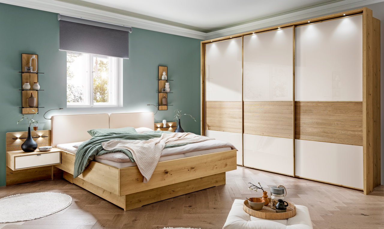 Moderner Schwebetürenschrank mit drei Türen in Holzoptik und weißen Paneelen, beleuchtet von oben. Perspektive: Frontalansicht im Schlafzimmer.