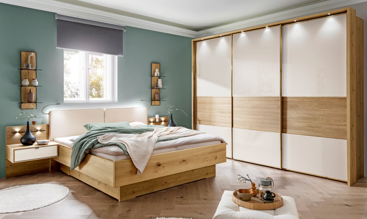 Moderner Schwebetürenschrank mit drei Türen in Holzoptik und weißen Paneelen, beleuchtet von oben. Perspektive: Frontalansicht im Schlafzimmer.