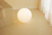 Leuchtende, kugelförmige Bodenlampe aus opalweißem Glas mit warmweißem Licht auf hellem Boden neben Sofa und Vorhang, aufgenommen aus leicht erhöhter Frontansicht.