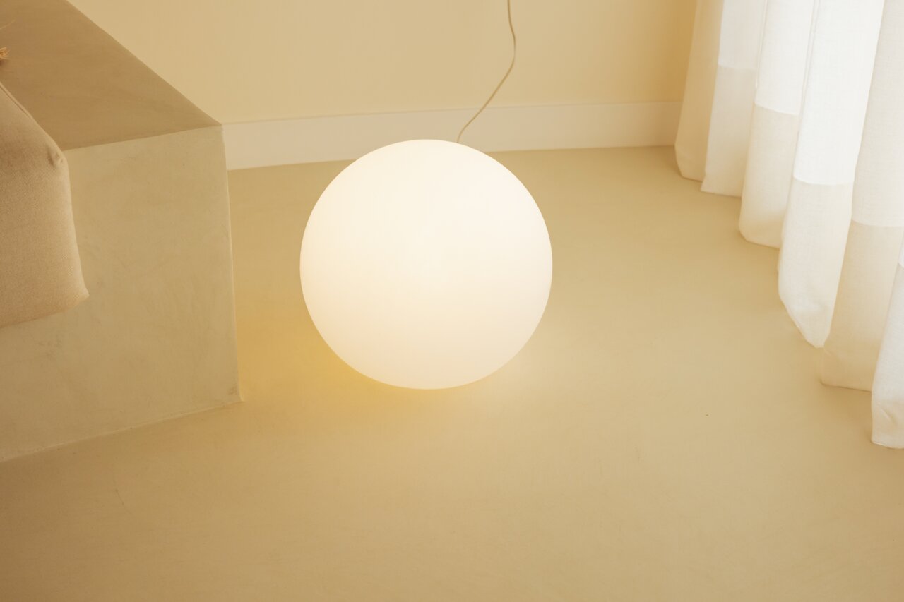 Een stralende, bolvormige staande lamp van opaalwit glas met warmwit licht op een lichte vloer naast een bank en een gordijn, gefotografeerd vanuit een licht verhoogd vooraanzicht.
