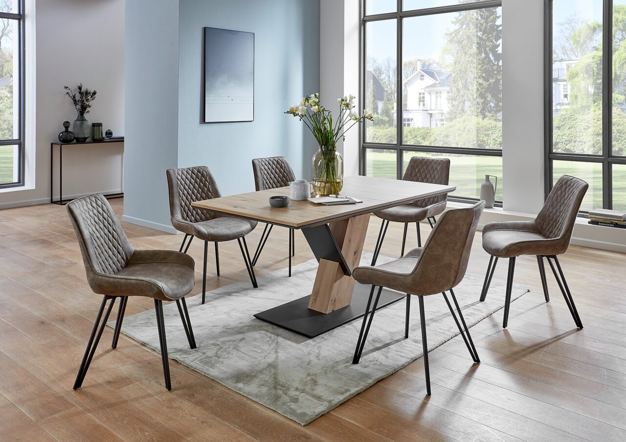 Moderner Esstisch mit Holzplatte und Metallgestell, umgeben von sechs gepolsterten Stühlen in einem hellen Raum mit großen Fenstern, seitliche Perspektive.