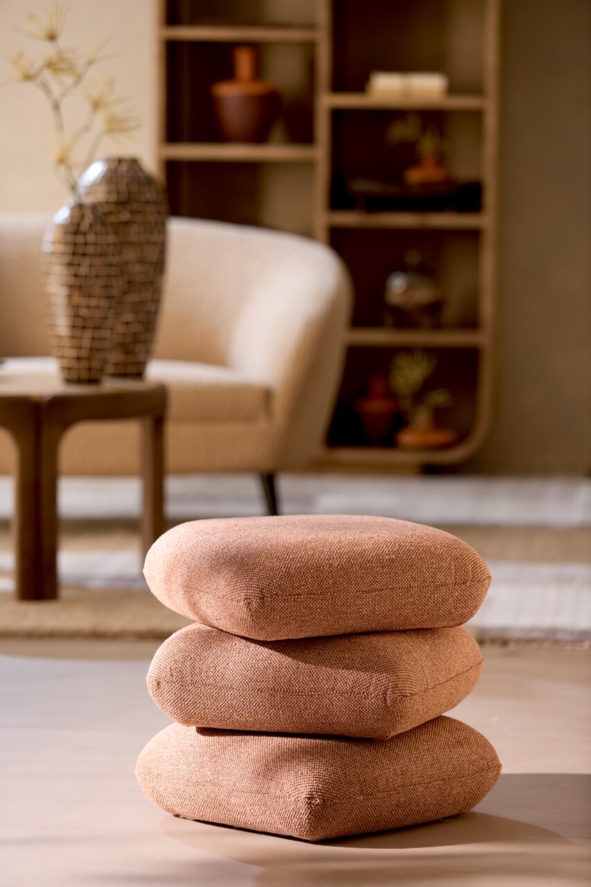 Ein Sitzpuff in warmem Braunton, bestehend aus drei übereinander gestapelten Kissen, in einem stilvoll eingerichteten Wohnzimmer. Perspektive: seitlich.