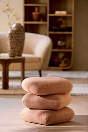 Ein Sitzpuff in warmem Braunton, bestehend aus drei übereinander gestapelten Kissen, in einem stilvoll eingerichteten Wohnzimmer. Perspektive: seitlich.