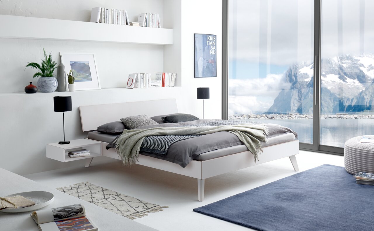 Modernes Bettgestell in einem hellen, stilvollen Schlafzimmer mit Blick auf eine Berglandschaft durch große Fenster, seitliche Perspektive.
