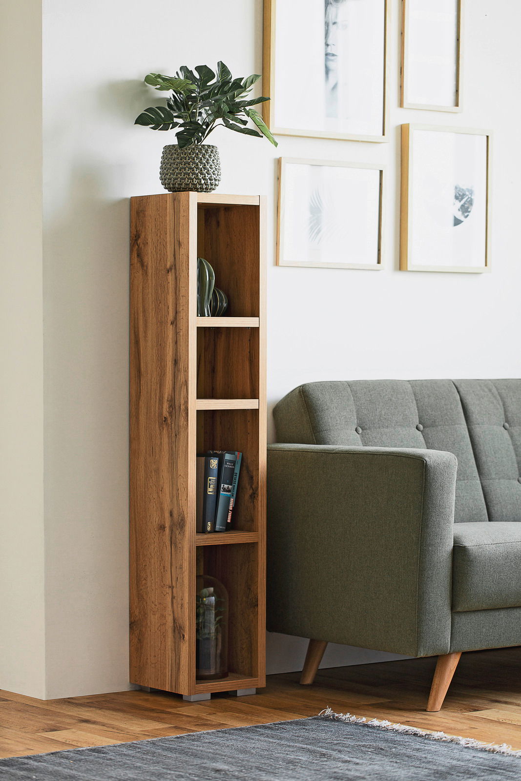 Schmales, hohes Regal aus Holz mit mehreren Fächern, dekoriert mit Büchern und Pflanzen, neben einem grauen Sofa in einem modernen Wohnzimmer. Perspektive von vorne.