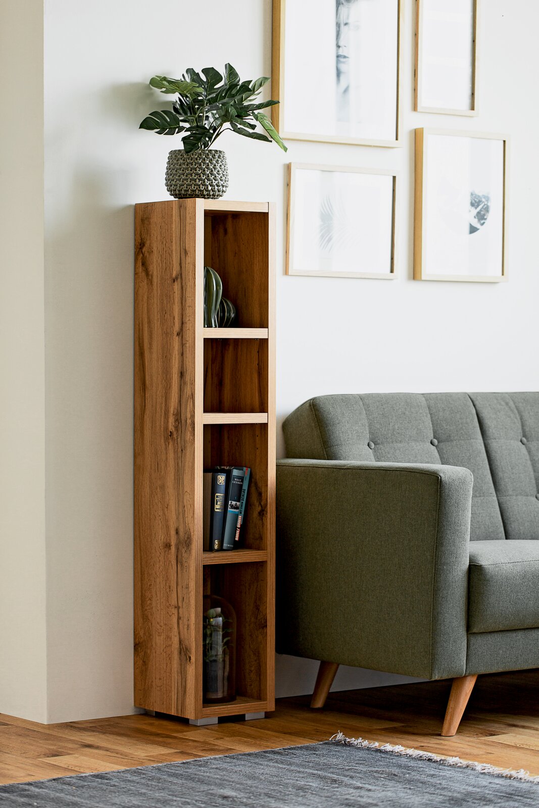 Een smalle, hoge houten kast met meerdere vakken, versierd met boeken en planten, naast een grijze bank in een moderne woonkamer. Aanzicht van voren.