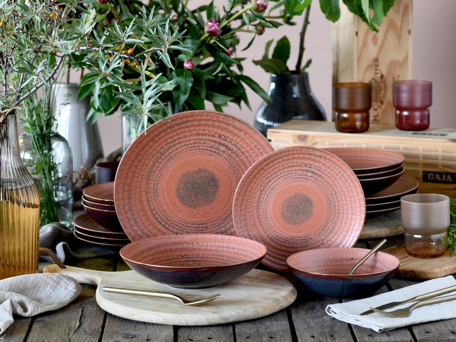 Rustikales Keramikgeschirr-Set in Rosé mit strukturierter, konzentrischer Oberfläche und dunklem Rand, bestehend aus Tellern und Schalen auf gedecktem Holztisch mit Besteck und Deko – Frontansicht in leicht schräger Perspektive.