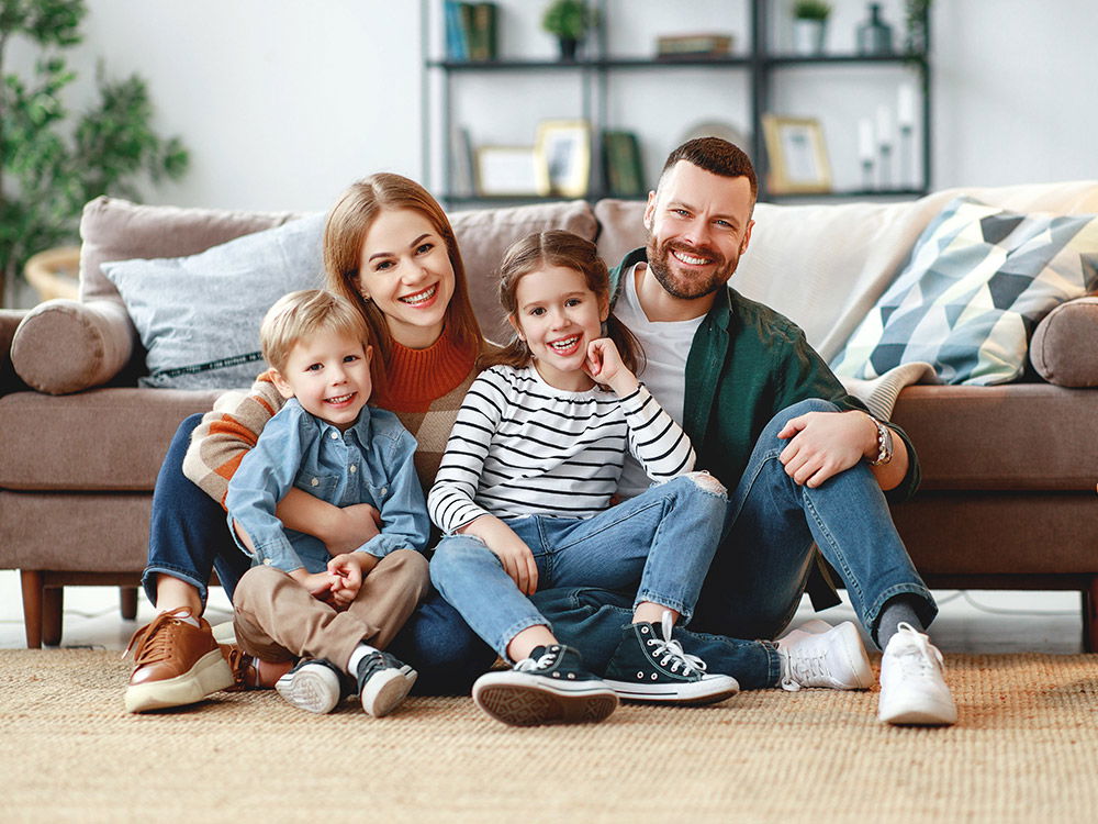 Familie sitzt zusammen auf dem Boden im Wohnzimmer vor einem braunen Sofa.