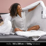 Frau sitzt lächelnd auf einem Bett und hält eine weiße Winterbettdecke mit den Maßen 155x220 cm, gefüllt mit 1260g Material, in den Händen. Die Decke hat schwarze Ränder und wird in einer gemütlichen Schlafzimmerumgebung präsentiert.