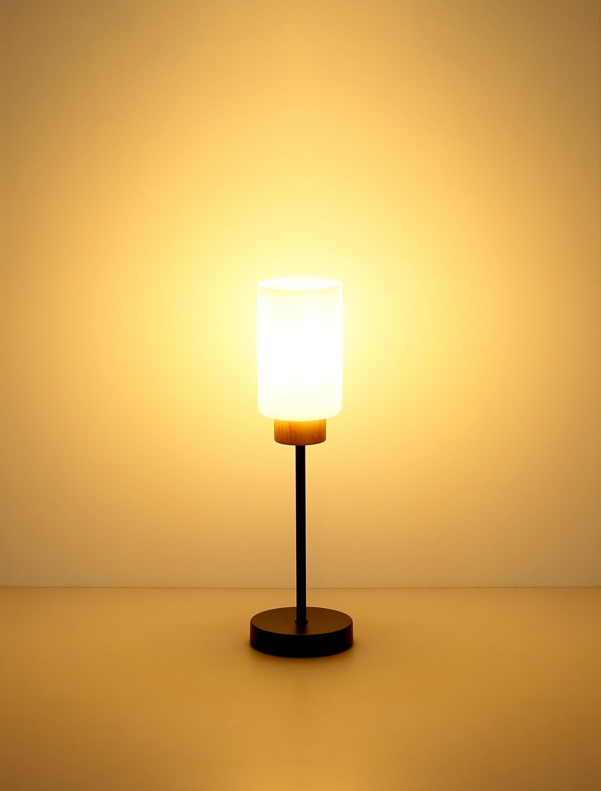 Moderne Tischlampe mit zylindrischem, weißem Glasschirm, schwarzem Stab und rundem Sockel mit Holzakzent, warm leuchtend vor neutralem Hintergrund, Frontalansicht