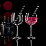 Zwei elegante Gin-Gläser mit Trinkhalmen, eines gefüllt mit rosa Getränk und Eiswürfeln, auf schwarzem Hintergrund mit unscharfen Flaschen im Hintergrund, seitliche Perspektive.