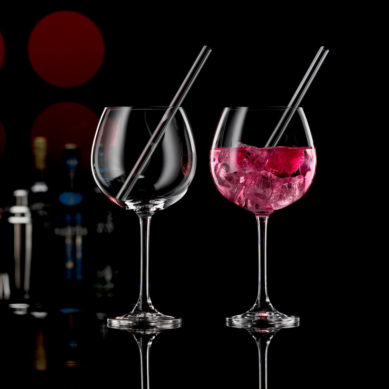 Novel Set van 2 gin-glazen met rietje Twee elegante gin-glazen met rietjes, waarvan één gevuld met een roze drankje en ijsblokjes, op een zwarte achtergrond met onscherpe flessen op de achtergrond, gezien vanuit een zijdelingse hoek.