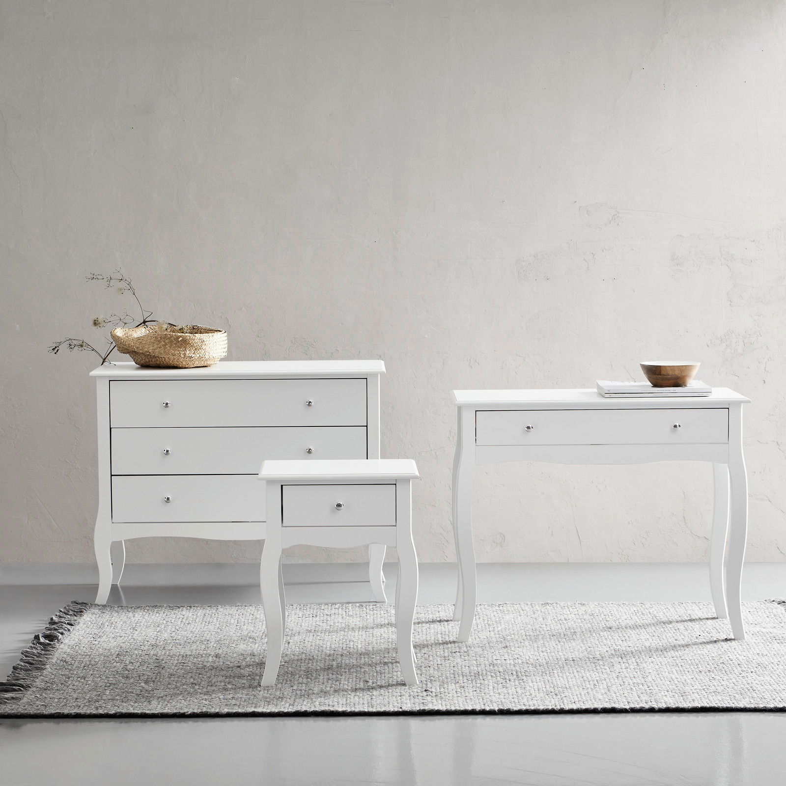 Drei weiße Möbelstücke auf einem Teppich vor einer hellen Wand, bestehend aus einer Kommode, einem Konsolentisch und einer Nachtkonsole, frontal betrachtet.