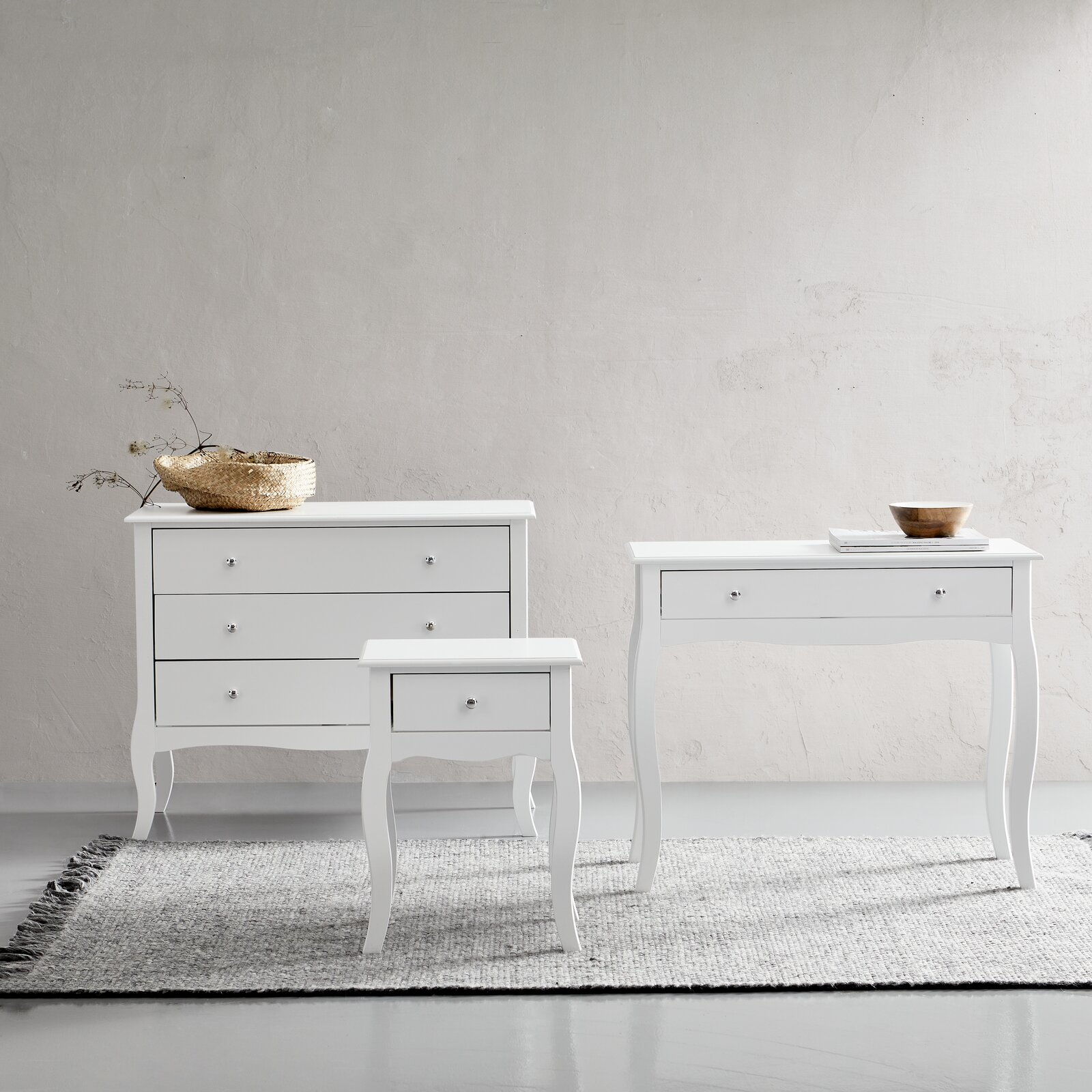 Drei weiße Möbelstücke auf einem Teppich vor einer hellen Wand, bestehend aus einer Kommode, einem Konsolentisch und einer Nachtkonsole, frontal betrachtet.