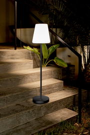 Schwarze, schlanke Stehleuchte mit weißem Lampenschirm auf einer Außentreppe aus Stein, warm leuchtend neben Pflanzen; Perspektive: frontale Ansicht auf Augenhöhe.