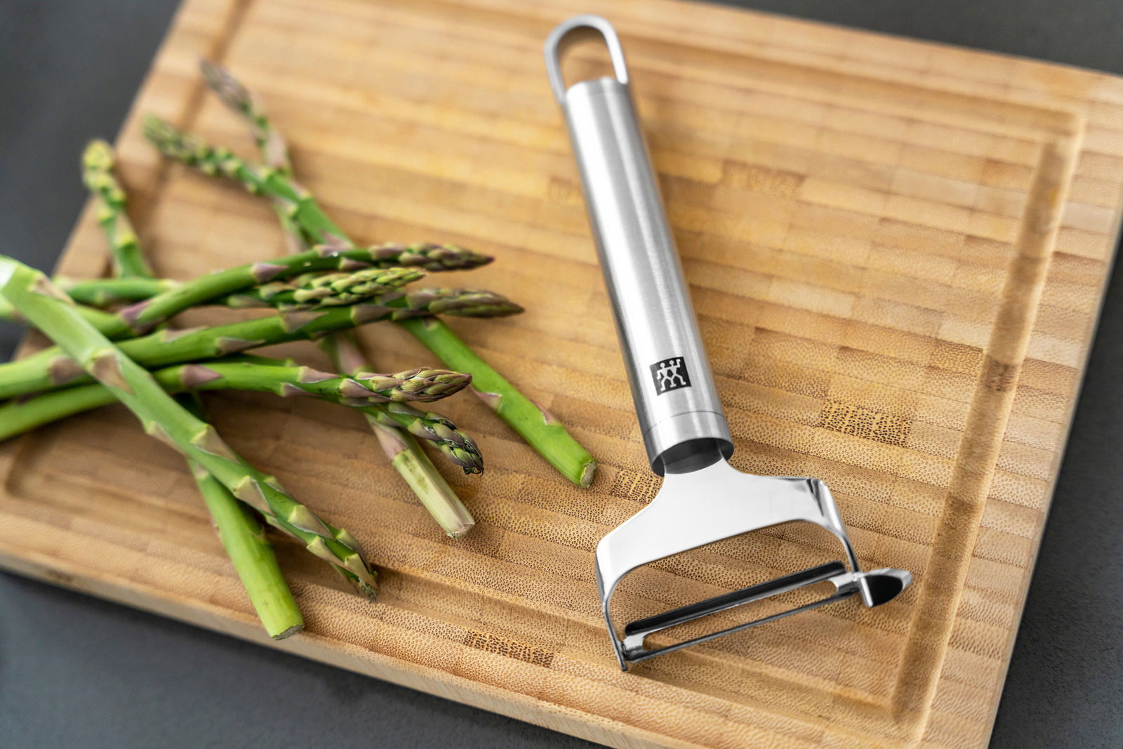 Pendelschäler quer Pro aus Edelstahl auf einem Holzbrett neben frischem grünem Spargel, von oben fotografiert.