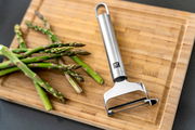 Pendelschäler quer Pro aus Edelstahl auf einem Holzbrett neben frischem grünem Spargel, von oben fotografiert.