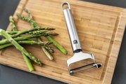 Pendelschäler quer Pro aus Edelstahl auf einem Holzbrett neben frischem grünem Spargel, von oben fotografiert.