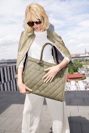 Frau mit Sonnenbrille hält eine olivgrüne, gesteppte Einkaufstasche mit zwei Henkeln auf einer Dachterrasse.