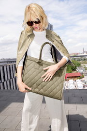 Frau mit Sonnenbrille hält eine olivgrüne, gesteppte Einkaufstasche mit zwei Henkeln auf einer Dachterrasse.