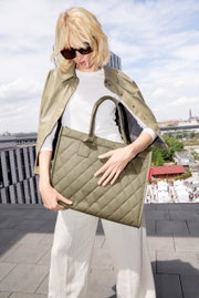 Frau mit Sonnenbrille hält eine olivgrüne, gesteppte Einkaufstasche mit zwei Henkeln auf einer Dachterrasse.