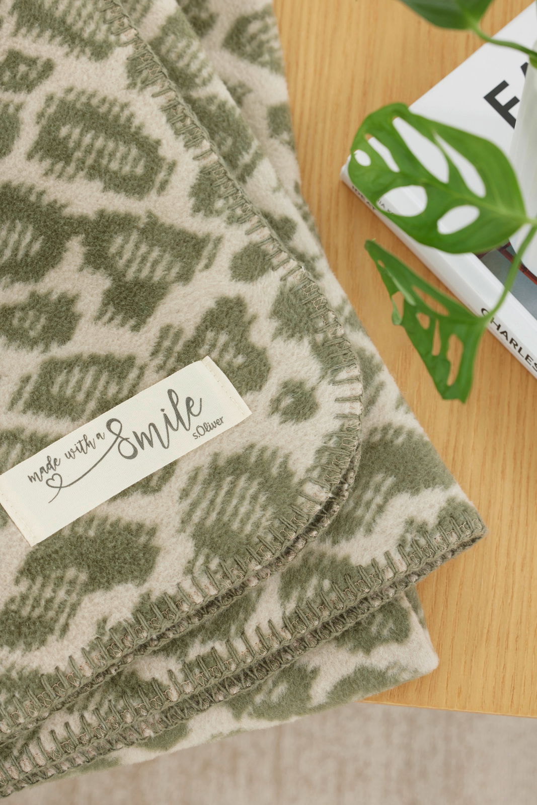 Nahaufnahme einer grünen und cremefarbenen Wohndecke mit geometrischem Muster und einem Etikett mit der Aufschrift 'made with a smile s.Oliver', auf einem Holztisch neben einem Buch und einer Pflanze.
