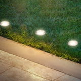 Bodenleuchten aus Edelstahl, eingebaut in eine Rasenfläche neben einem gepflasterten Weg, aus der Vogelperspektive betrachtet.