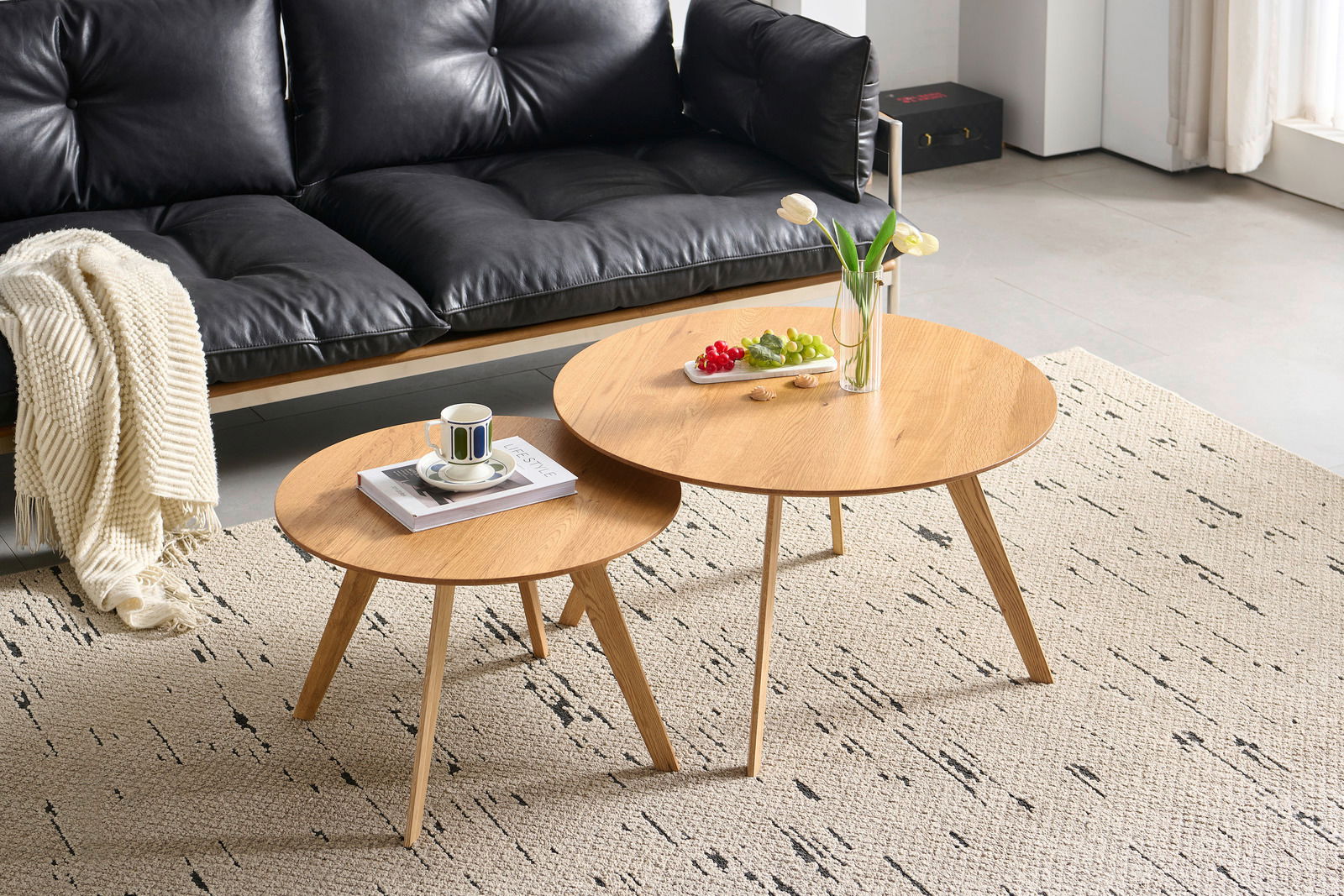 Z2 Salontafel (set) MEAGAN Zwei runde Couchtische aus Holz in einem modernen Wohnzimmer, fotografiert aus einer leicht erhöhten Perspektive. Auf den Tischen stehen eine Vase mit Blumen, ein Teller mit Obst und eine Tasse auf einem Buch.