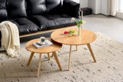 Zwei runde Couchtische aus Holz in einem modernen Wohnzimmer, fotografiert aus einer leicht erhöhten Perspektive. Auf den Tischen stehen eine Vase mit Blumen, ein Teller mit Obst und eine Tasse auf einem Buch.