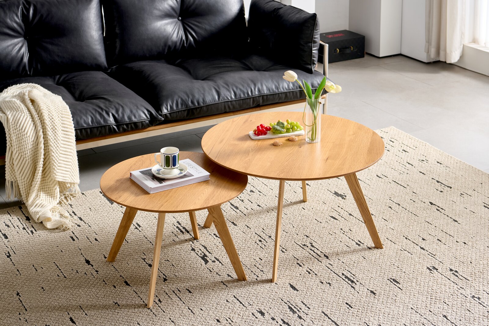 Z2 Couchtisch (Set) MEAGAN Zwei runde Couchtische aus Holz in einem modernen Wohnzimmer, fotografiert aus einer leicht erhöhten Perspektive. Auf den Tischen stehen eine Vase mit Blumen, ein Teller mit Obst und eine Tasse auf einem Buch.