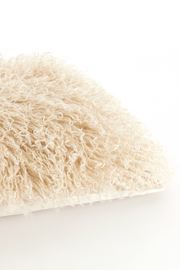 Nahaufnahme eines cremefarbenen, lockigen Fell-Kissens auf weißem Hintergrund, schräg von oben fotografiert.