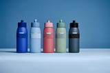 Fünf Sporttrinkflaschen in verschiedenen Farben (blau, hellblau, rosa, grün, grau) in einer frontalen Perspektive auf einem blauen Hintergrund.