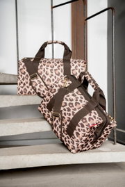 Zwei Reisetaschen mit Leopardenmuster, eine auf einer Treppe liegend und die andere aufrecht stehend, mit braunen Tragegurten und goldenen Schnallen, seitliche Perspektive.