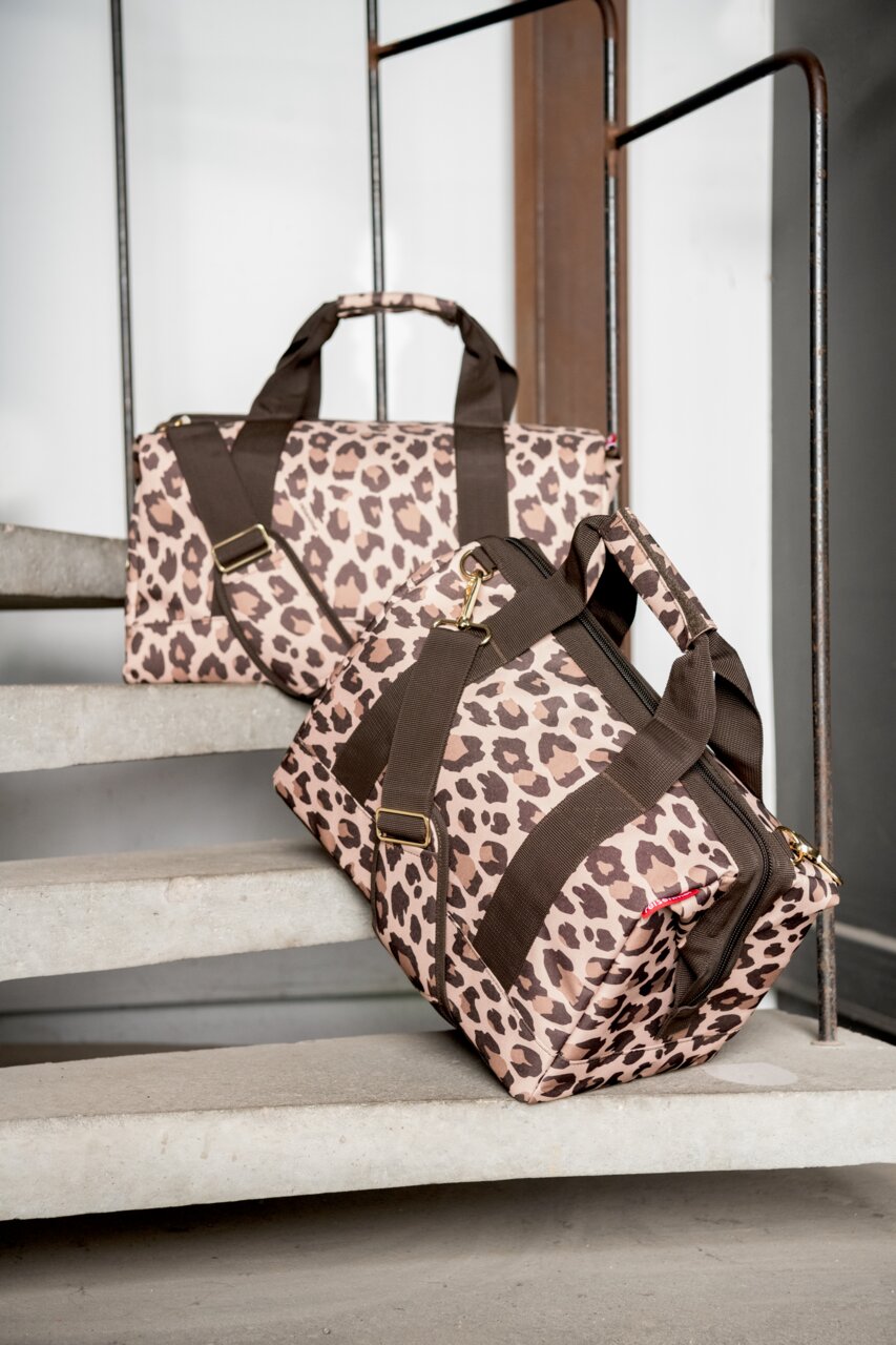 Zwei Reisetaschen mit Leopardenmuster, eine auf einer Treppe liegend und die andere aufrecht stehend, mit braunen Tragegurten und goldenen Schnallen, seitliche Perspektive.