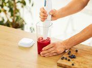 Person benutzt einen weißen Stabmixer in einem transparenten Messbecher mit rotem Inhalt, auf einem Holztisch mit Blaubeeren. Perspektive von oben.