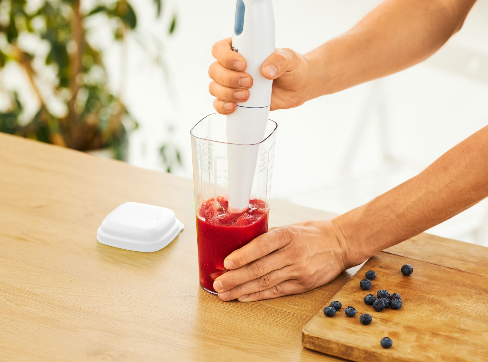 Person benutzt einen weißen Stabmixer in einem transparenten Messbecher mit rotem Inhalt, auf einem Holztisch mit Blaubeeren. Perspektive von oben.