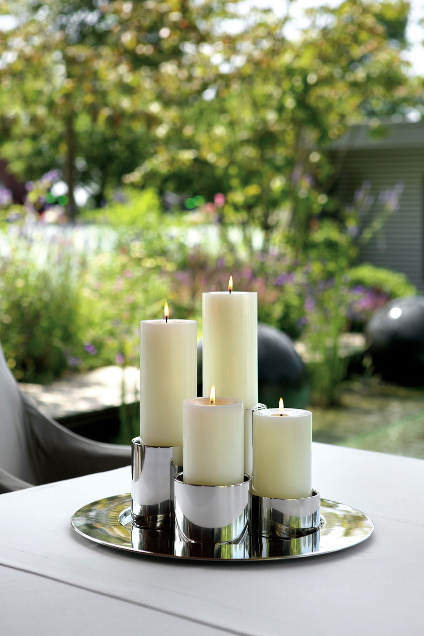 Eleganter Edelstahl-Stumpenhalter mit brennenden Kerzen, auf einem Tisch im Freien platziert, umgeben von einem grünen, unscharfen Gartenhintergrund. Perspektive von schräg oben.