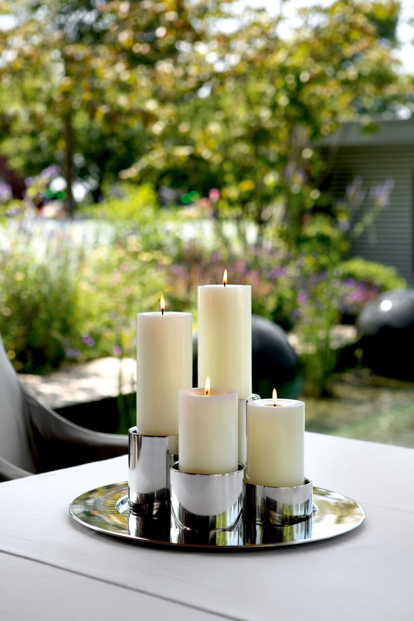 Elegante roestvrijstalen kaarsenhouder met brandende kaarsen, geplaatst op een tafel in de open lucht, omgeven door een groene, wazige tuinachtergrond. Perspectief vanuit een schuine hoek van bovenaf.