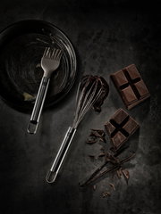 Kleiner Schneebesen aus Edelstahl, von oben fotografiert, neben einer Pfanne mit Backpinsel und Schokoladenstücken.