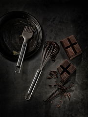 Kleiner Schneebesen aus Edelstahl, von oben fotografiert, neben einer Pfanne mit Backpinsel und Schokoladenstücken.