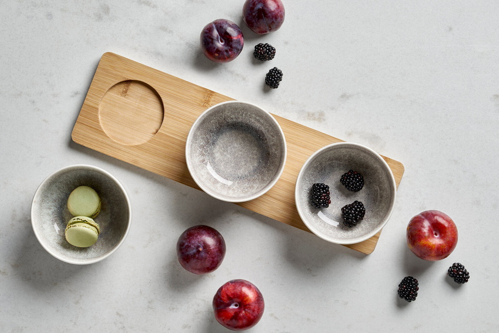 Vogelperspektive auf ein 4-teiliges Schalen-Set mit einem Holztablett, gefüllt mit Macarons, Brombeeren und Pflaumen auf einem hellen Untergrund.