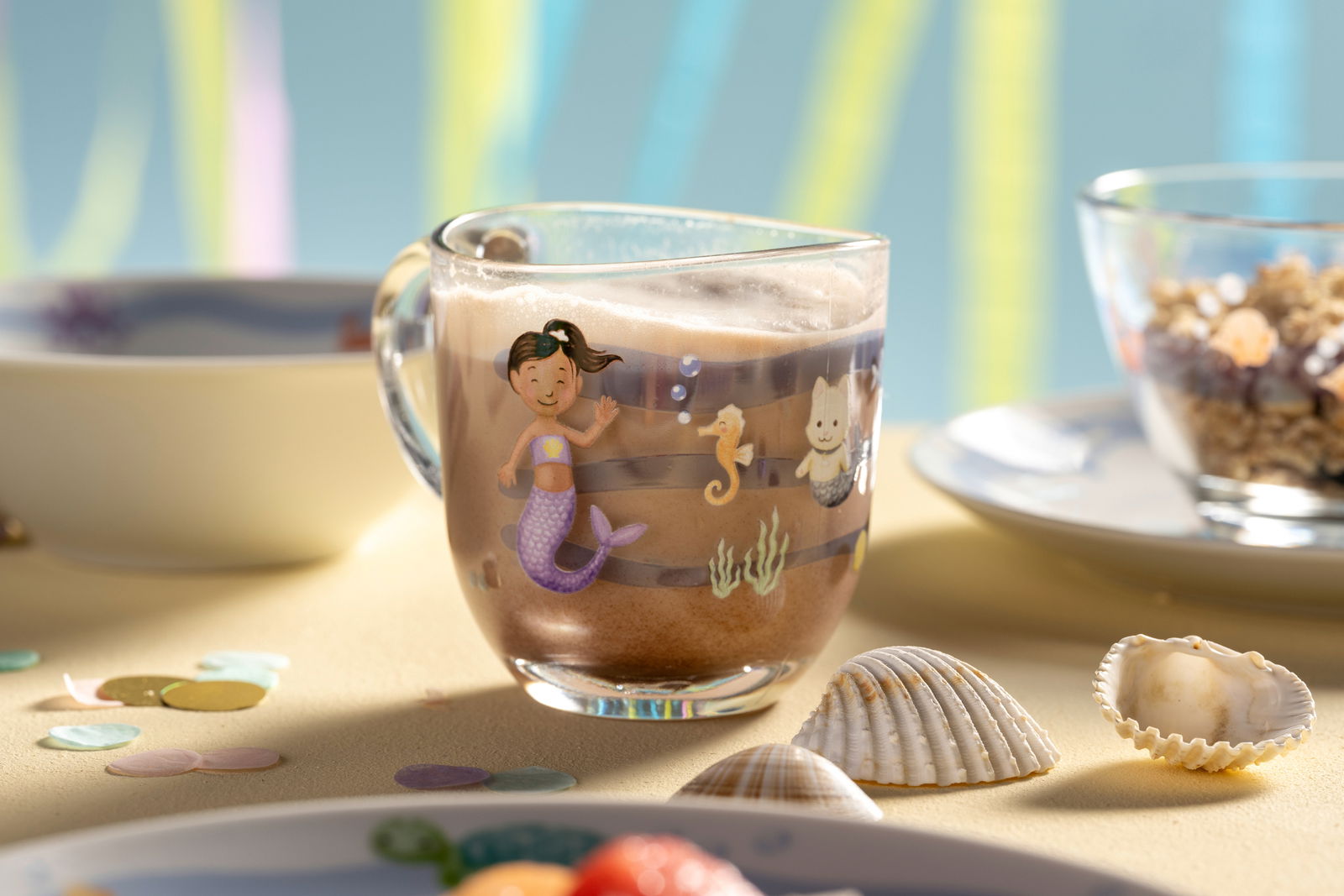 Kindertasse mit Meerjungfrauenmotiv, gefüllt mit Kakao, auf einem Tisch mit Muscheln und Konfetti, seitliche Perspektive