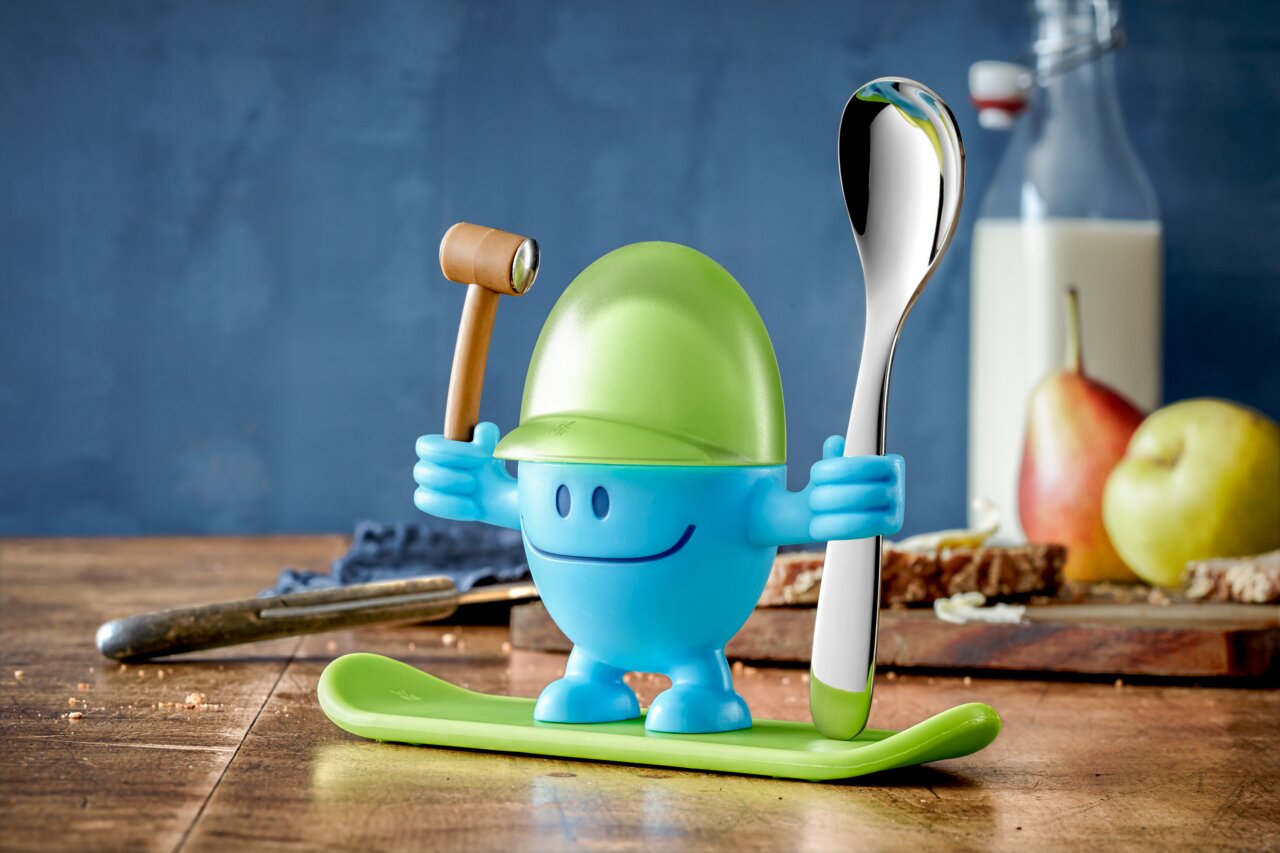 Blauer Eierbecher McEgg mit grünem Helm, Löffel und Hammer, auf einem Tisch mit Frühstückszubehör, seitliche Perspektive