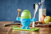 Blauer Eierbecher McEgg mit grünem Helm, Löffel und Hammer, auf einem Tisch mit Frühstückszubehör, seitliche Perspektive