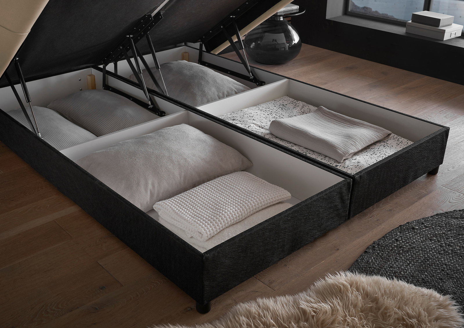 Polsterbett mit geöffnetem Bettkasten, gefüllt mit Kissen und Decken, aus der Vogelperspektive fotografiert.